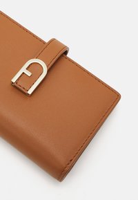 Billetera de cuero marrón con una textura suave, que presenta un cierre de metal dorado con forma de "A" y un detalle de borde cosido.