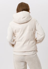 Crèmekleurige, gewatteerde jas met een capuchon met hoge hals. Heeft een zachte, gladde stof met een getailleerd silhouet en minimale stiksel details.