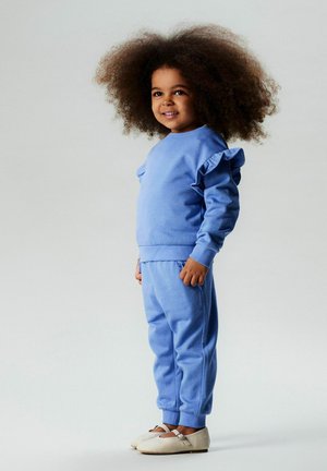 Vaaleansininen lasten collegepaita, jossa on röyhelöityjä olkapääyksityiskohtia, yhdistettynä vastaaviin verkkarihousuihin. Pehmeä kangas, rento istuvuus ja joustavat hihansuut.