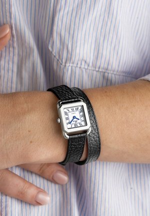 Ranne, jossa on musta kaksinkertainen nahkaranneke, suorakaiteen muotoinen hopeinen kotelo ja valkoinen kellotaulu sinisillä osoittimilla ja roomalaisilla numeroilla.