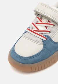 Blå mokka og hvit skinn-sneaker med borrelås, rød og hvit stripete snøring, og en teksturert gummisåle.