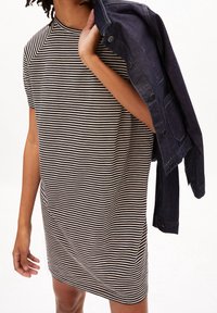 Osoba ubrana w sukienkę w czarno-białe paski z krótkim rękawem, trzymająca ciemnoniebieską dżinsową kurtkę na ramieniu.