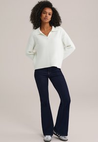 Suéter blanco acanalado con cuello, combinado con unos vaqueros de campana azul oscuro y zapatillas grises. El conjunto presenta un ajuste relajado y un estilo casual.