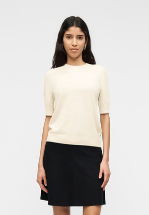 Donna con lunghi capelli scuri, indossa una maglietta crema a maniche corte e una gonna nera al ginocchio, in piedi davanti a uno sfondo grigio chiaro.