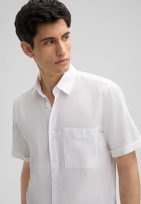 Kurzärmeliges weißes Shirt aus strukturiertem Stoff, mit Knopfleiste vorne und einer Brusttasche. Einfacher Kragen und hochgekrempelte Ärmel.