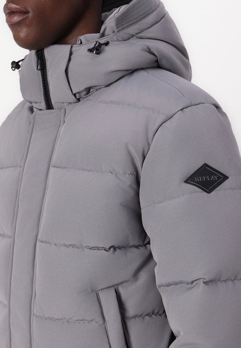 Acercamiento de una persona que lleva una chaqueta acolchada ligera de color gris con capucha y un parche del logo de Replay en la manga.