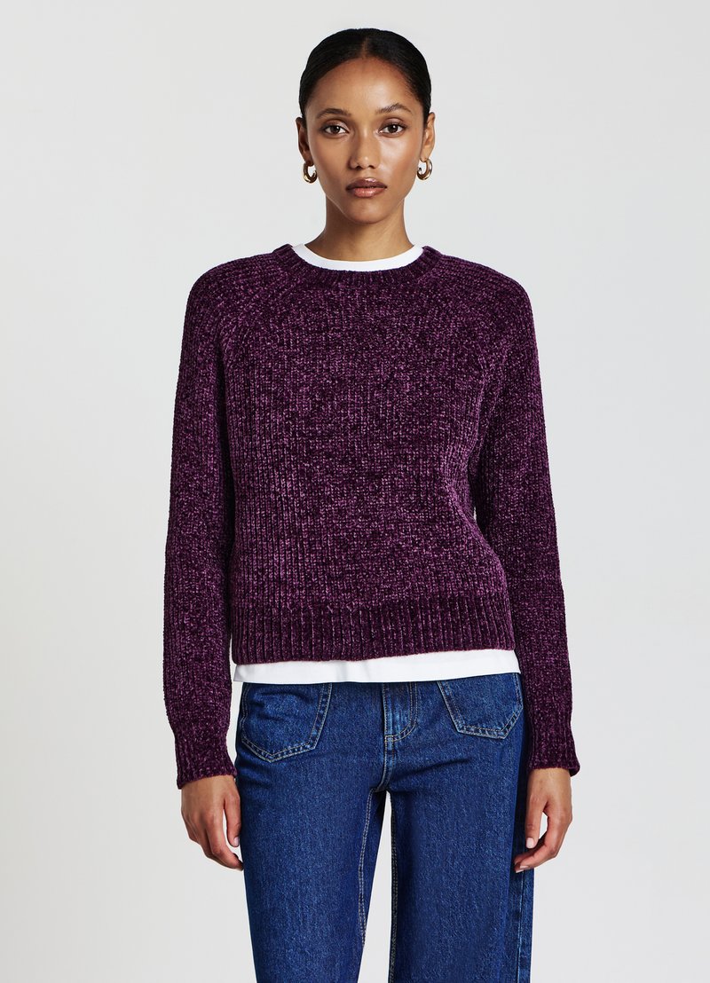 Lila gestrickter Pullover mit strukturiertem Muster, Rundhalsausschnitt und gerippten Bündchen. Über einem weißen Hemd getragen, kombiniert mit blauen Jeans.