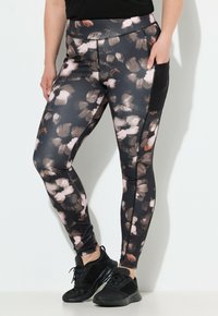 Donna con leggings neri decorati con motivi floreali astratti marroni e beige, tasca laterale in rete e scarpe da ginnastica nere.