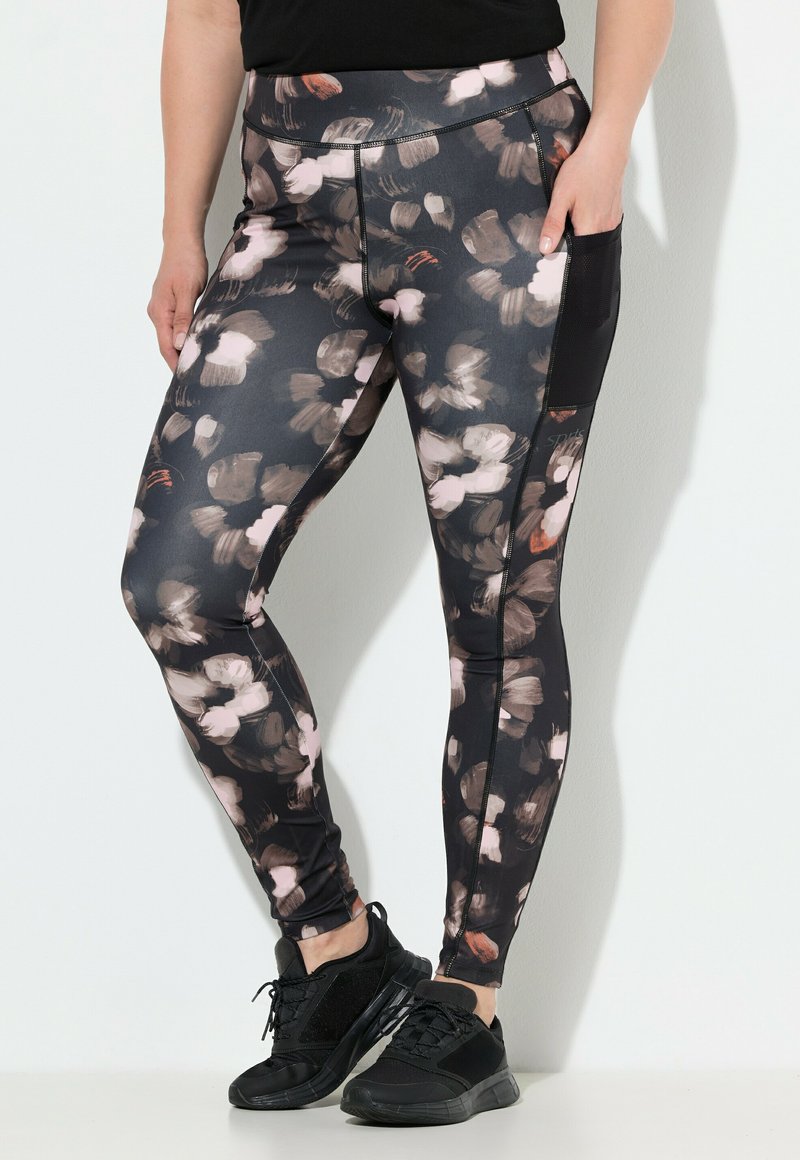 Donna con leggings neri decorati con motivi floreali astratti marroni e beige, tasca laterale in rete e scarpe da ginnastica nere.