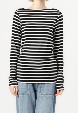 Osoba ubrana w czarnobiały, poziomo prążkowany, długorękawowy sweter, zestawiony z jasnoniebieskimi dżinsami, obciętymi na szyi i na wysokości bioder.