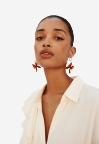 Un paio di grandi orecchini a forma di farfalla color ambra con hardware dorato, indossati da una modella in una blusa color crema con dettagli arricciati.