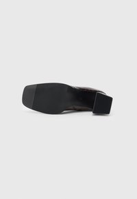 Suela de un zapato marrón oscuro con un tacón cuadrado, suela de goma texturizada y una sección superior ligeramente decorada visible.