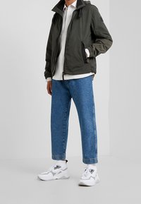 Olive green lightweight jacket with a zip, hood, and side pockets, paired with light blue straight-leg jeans and white sneakers.