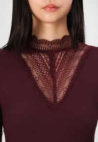 Burgundy-huulinen toppi, jossa on korkea pitsikaulus, jossa on aaltoilevat reunat ja monimutkaiset kukkakuviot. Rypistynyt pinta kauttaaltaan.