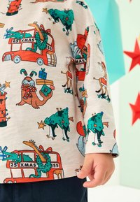 Chemise à manches longues en tissu crème ornée de motifs de Noël en cartoon colorés, incluant des animaux, des arbres et des cadeaux. Présente un ourlet arrondi.