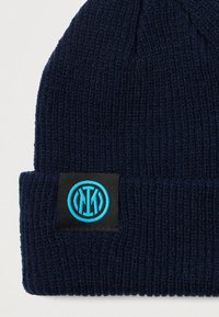 Gorro de malha azul-marinho com um tecido texturizado, apresentando um patch preto com um logótipo circular azul. Design com aba dobrada.