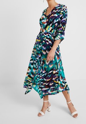 Femme portant une robe midi à motif abstrait coloré avec un col en V, des manches trois-quarts et des sandales à talons blocs blanches avec bride à la cheville.