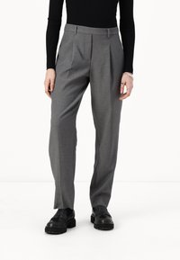 Pantalons gris ajustés avec des plis, présentant une texture lisse et un design droit. Portés avec des chaussures noires.