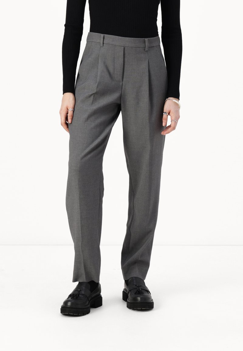 Pantalons gris ajustés avec des plis, présentant une texture lisse et un design droit. Portés avec des chaussures noires.
