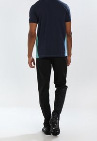 Man i svarta byxor, svarta skor och en marinblå skjorta med gröna och blå sidopaneler som går bort från en vit bakgrund.