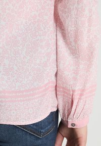 Blusa rosa chiaro con motivo floreale e texture trasparente. Presenta maniche arricciate, un dettaglio con bottoni e una fascia decorativa all'orlo.
