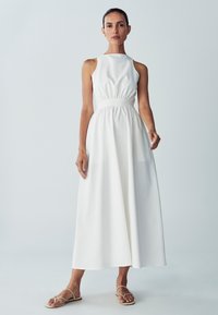 Weißes ärmelloses Kleid mit hohem Kragen, geraffter Taille und einem fließenden, knöchellangen Rock. Der Stoff wirkt glatt und leicht.