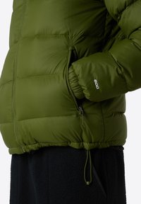 Närbild av en person som bär en olivgrön puffjacka med handen i en zip-försedd sidoficka och "600" broderat på ärmen.