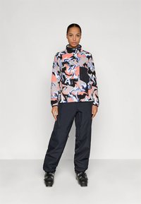 Meerlagige jas met abstract golvend patroon in roze, blauw en zwart; voorzien van een ritssluiting en een hoge kraag; gecombineerd met zwarte broek.