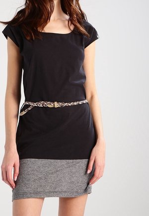 Woman wearing a short black and gray dress with a braided belt standing against a plain light background.