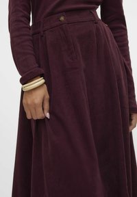 Burgundy corduroy-mekko, jossa on pitkät hihat, laskostettu hame ja nappiyksityiskohta vyötäröllä. Kädessä vaaleansävyisiä rannekoruja.