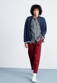 Navy track jacket with striped shirt, burgundy pants, and white sneakers; features a sporty design and relaxed fit, with contrasting stripes.
