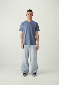 Jovem de pé, virado para a frente, vestindo uma t-shirt azul, jeans leves e largas, e sapatilhas pretas, contra um fundo claro e liso.