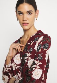 Maroon floral blouse with long sleeves, featuring cream and black flowers, deep V-neck, and a smooth fabric texture. Elegant design details.