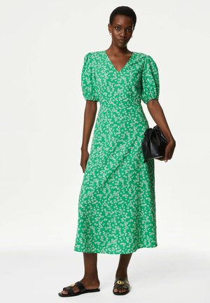 Groene bloemen midi-jurk met korte pofmouwen en een V-hals. Kenmerkt zich door witte madeliefje-achtige patronen en een vloeiende silhouette. Gecombineerd met een zwarte tas.