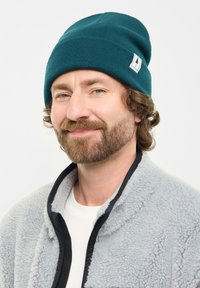 Teal knit beanie with a ribbed texture, featuring a white tag with a tree design. Worn with a gray fleece jacket and a white shirt.