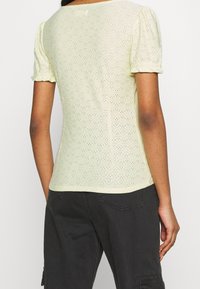 Nainen, jolla on päällään vaaleankeltainen lyhythihainen verkkopaita, jossa on puffihihat, ja mustat cargo-housut, näkyy selkäpuolelta.