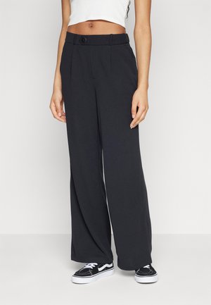 Pantaloni neri a gamba larga con una vestibilità sartoriale, chiusura con bottone e pieghe frontali. Indossati con sneakers nere. Tessuto dalla texture liscia.