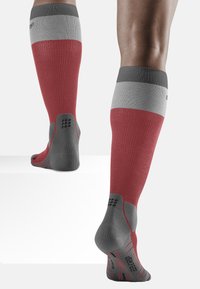 Calze compressive con un design a gradiente, con una sezione inferiore rossa, una fascia superiore grigia e dettagli testurizzati grigi sui talloni e sulle punte.