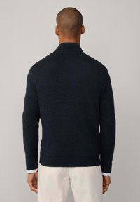 Dunkelblauer Strickpullover mit hohem Kragen und gerippten Bündchen, mit strukturierter Oberfläche und schlanker Passform, getragen über hellen Hosen.