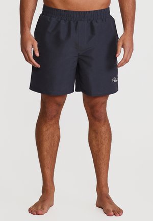 Mann iført marineblå shorts med elastisk midje, stående barbeint mot en ensfarget bakgrunn.