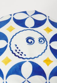 Tekstil i hvid med et blåt og gult geometrisk mønster, der indeholder cirkulære og diamantformede figurer samt designelementer, der ligner et ansigt.