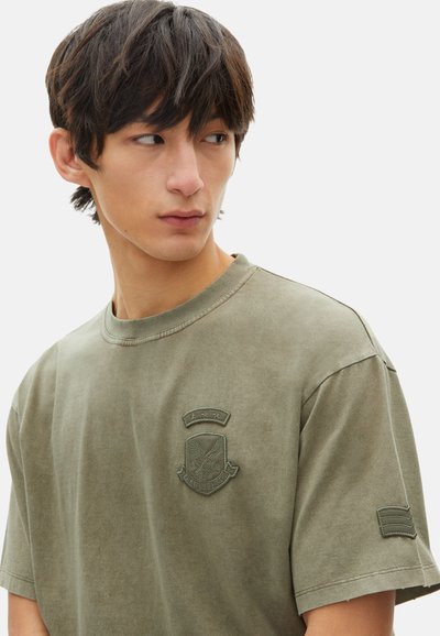 Camiseta de algodón verde oliva con escote redondo, mangas cortas y un parche texturizado que presenta un emblema en el pecho y el brazo.
