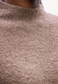 Gros plan d'une personne à la peau foncée portant un pull en laine douce et moelleuse de couleur marron clair, texturé, avec un col rond haut.