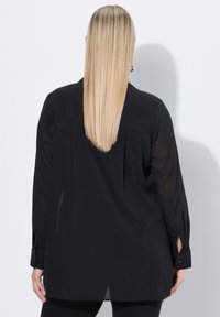 Schwarzes Langarmshirt mit lockerer Passform, das einen gefalteten Rücken, knöpfbare Manschetten und eine leicht transparente Textur aufweist.