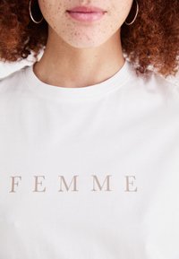 Bílý bavlněný tričko s nápisem "FEMME" v béžových velkých písmenech. Kolečkový výstřih a krátké rukávy. Hladký povrch.