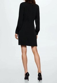 Frau in einem langärmligen schwarzen Kleid und schwarzen High Heels, von hinten vor einem schlichten weißen Hintergrund gezeigt.
