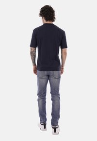 Uomo con capelli ricci, di spalle, indossa una maglietta nera, jeans blu, sneakers bianche e mostra un tatuaggio a tutto braccio sul braccio sinistro.