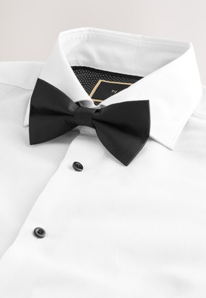 Camisa blanca de vestir con botones negros y pajarita negra con textura centrada bajo un cuello abierto.