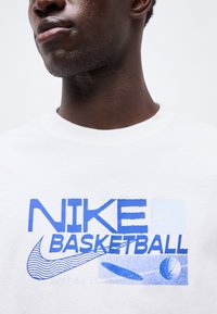 Κοντινή λήψη ενός ατόμου που φοράει λευκό πουκάμισο με μπλε κείμενο "NIKE BASKETBALL", κυματιστό λογότυπο Nike και γραφικά καλαθοσφαίρισης.