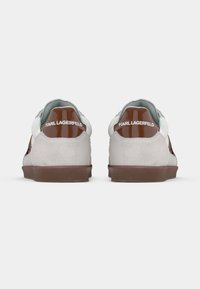 Grå og hvite semskede joggesko med blanke brune hældetaljer og merkevaremerking. Flate brune gummisåler, rund form og teksturert overflate.